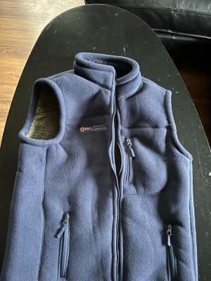 Blå fleeceväst från Norway - Mysig blå fleeceväst från Norway med hög krage och dragkedja framtill. Västen har tre fickor med dragkedja och ett mjukt, beige teddyfoder på insidan. Perfekt för lager-på-lager och outdoor-vibes.