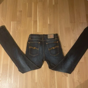 Mörkblå slim jeans från Nudie Jeans - ((W29L32)) Säljer ett par mörkblå slim jeans från Nudie Jeans med klassisk orange kontrastsöm och vågig brodyr på bakfickorna. Jeansen har dragkedja, fem fickor och är tillverkade i slitstarkt jeansmaterial. Perfekta för dig som gillar en smal passform och stilren look.