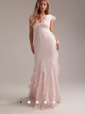 Ljusrosa balklänning - Superfin ljusrosa långklänning med v-ringning och volangdetaljer längs axlar och kjol. Klänningen har ärmlös design med breda axelband och en öppen rygg. Klänning är helt ny med prislapp kvar och säljer pga att den ej passade på mig och hann inte skicka tillbaka, nypris är 1500kr