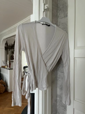 Vit/Beige omlottopp från 157 - Säljer en vit/beige omlottopp från 157 i mjukt material. Toppen har långa ärmar, v-ringning och knytband i midjan som ger en snygg siluett. Perfekt att styla med jeans eller kjol för en enkel men chic look.