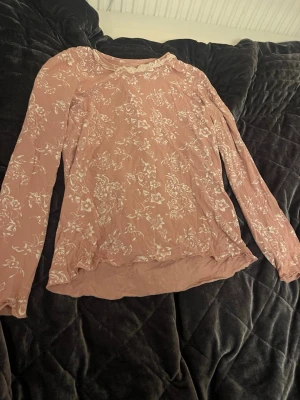 Rosa blommig pyjama sett med spetsdetalj - Söt rosa pyjamas med vitt blommönster, rosett och spetskant vid halsen. Pyjamasen har en mjuk och tunn känsla och är i storlek 134-140. Funderingar? Skriv till mig💕