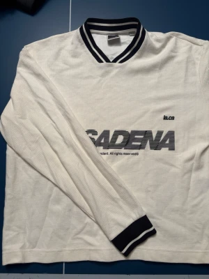 Vit långärmad pikétröja Jack & Jones - Säljer en vit långärmad tröja från Jack & Jones Core med svart texttryck 'PASADENA' framtill. Tröjan har svartvitrandig krage och muddar, samt klassisk stil. Perfekt för dig som gillar sportig och clean stil. Lite cropped vid midjan. Skriv om funderingar 🙌