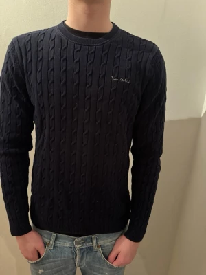 Mörkblå kabelstickad tröja BondeLid - Säljer en mörkblå kabelstickad tröja från BondeLid. Tröjan har rund halsringning, diskret broderad logga på bröstet och långa ärmar. Klassisk och stilren design som passar perfekt till jeans.