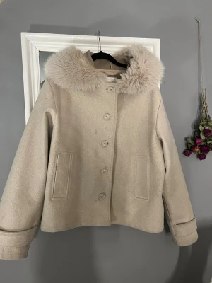 Beige kappa med fuskpäls från Gina Tricot - Snygg beige kappa från Gina Tricot med stor fluffig fuskpäls-krage och raka fickor. Jackan har knappar framtill. Perfekt för dig som vill ha en stilren och trendig look i höst och vinter.