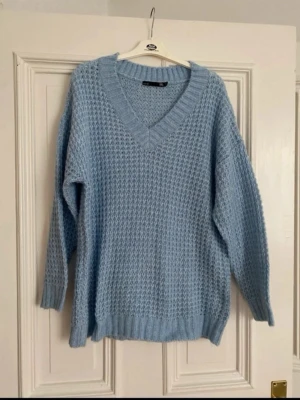 Ljusblå stickad tröja från 157 - Mysig ljusblå stickad tröja från 157 med v-ringad hals och grovstickat mönster. Tröjan har lång ärm och ribbade muddar vid ärmslut och nederkant. Perfekt för lager på lager under kyliga dagar.