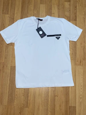 Vit t-shirt  med logga - Snygg vit t-shirt  med svart logotypdetalj och en ikonisk trekantig patch på bröstet. Klassisk rund hals och korta ärmar. Tillverkad i mjuk bomull för skön känsla och stilren look.