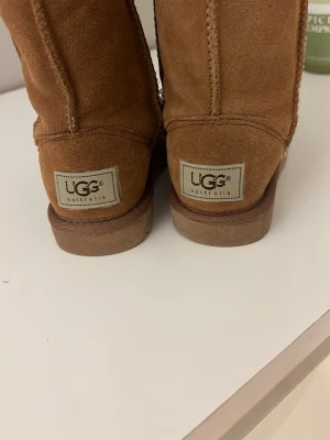 Bruna UGG boots i mocka - Klassiska bruna UGG boots i mjuk mocka med fluffigt foder och rund tå. Snyggt broderade sömmar och rejäl platt sula för extra komfort. Perfekta för kalla dagar och ger en avslappnad vibe till din outfit.