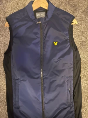 Blå dunväst från Lyle & Scott - Säljer en snygg marinblå dunväst från Lyle & Scott med gul logga på bröstet. Västen har svarta sidopaneler, hög krage och dragkedja framtill. Perfekt att slänga över en hoodie eller tröja för extra värme.