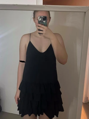 Svart volangklänning med smala axelband - Supersöt svart kort klänning med smala axelband och v-ringning. Funkar som myskläbning och finklänning. 