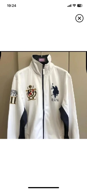Vit ziptröja / jacka från US Polo Assn - Säljer en vit vindjacka från US Polo Assn med marinblå detaljer och broderade loggor på bröstet och ärmen. Jackan har hög krage, dragkedja framtill och sportig look. Perfekt för dig som gillar preppy stil och vill sticka ut med snygga emblem.