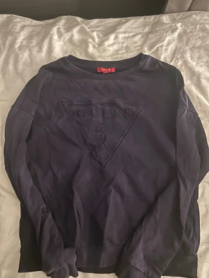 Mörkblå sweatshirt från Guess - Säljer en mörkblå sweatshirt från Guess med det klassiska triangellogotypen ton-i-ton på bröstet. Tröjan har rund halsringning och långa ärmar. Perfekt för dig som gillar stilrena och enkla plagg med en twist.