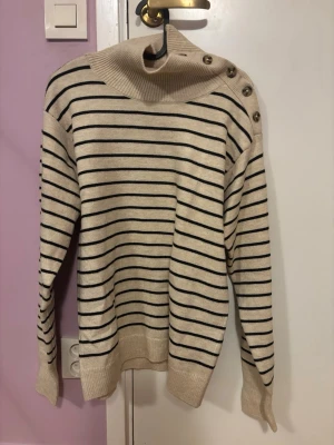 Randig beige polotröja från Soaked - Säljer en beige polotröja från Soaked i storlek XS. Tröjan har svarta horisontella ränder, hög krage med dekorativa knappar på axeln och långa ärmar. Materialet är mjukt och stickat, perfekt för kyligare dagar.