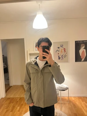 Ljusgrön regnjacka från RAINS - Säljer en stilren ljusgrön regnjacka med huva och dragkedja framtill. Jackan har två stora bröstfickor med lock och justerbar huva med snörning. Perfekt för regniga dagar och enkel att matcha med olika outfits. Ordinarie pris 2750kr. 