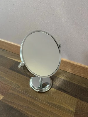 Rund sminkspegel  - Säljer en stilren rund sminkspegel med silverfärgad ram och fot. Spegeln har justerbar vinkel och står stadigt på ett bord eller sminkbord. Tror materialet är i hårt plast 