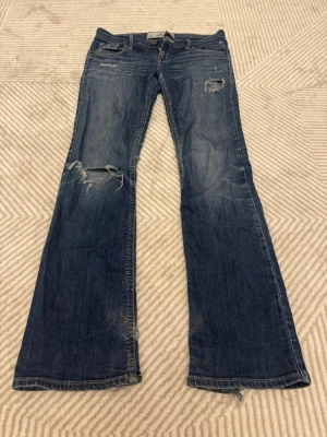Blå bootcut jeans från hollister - Säljer ett par blå jeans med bootcut passform och slitna detaljer på knän och bakfickor. Jeansen har klassisk femficksdesign, låg midja och är tillverkade i ett mjukt denimtyg. Perfekta för en avslappnad och trendig look.