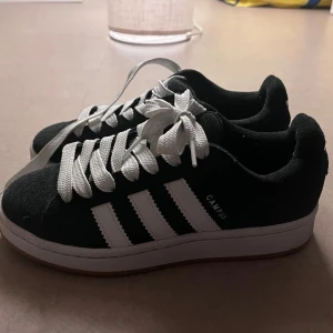 Svarta Adidas Campus sneakers - Säljer ett par klassiska Adidas Campus sneakers i svart mocka med vita detaljer och de ikoniska tre ränderna på sidan. Skorna har knappt kommit till användning och därför är fortfarande ganska fräscha och ser ut som helt nya. Strl 38 men passar nog den som har strl 37.5 eller 37 då jag har strl 37 i andra skor. Kommer med svarta snören då jag fick både svarta snören och vita när jag köpte skorna.