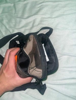 Camouflage axelväska från Adidas - Säljer en liten och smidig axelväska från Adidas med camouflage-mönster och orange logga framtill. Väskan har justerbar svart rem, dragkedja och flera fack inuti. Perfekt för att bära med sig det viktigaste och har en cool streetstil.