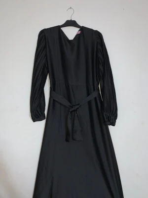 Svart långklänning från La Pablo - Snygg svart långklänning från La Pablo med plisserade långärmar och knytband i midjan. Klänningen har en mjuk satinliknande yta och en enkel rund halsringning. Perfekt för dig som gillar stilrena och eleganta plagg med coola detaljer.