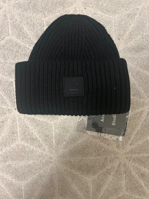  Acne Studios Mössa - Svart ribbstickad mössa från Acne Studios med ikonisk face patch framtill. Tillverkad i 100% ull och har en bred uppvikt kant. Perfekt för att hålla dig varm och ge din look en clean vibe.