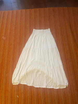 Vit långkjol från 157, storlek M - Säljer en vit långkjol från 157 i storlek M men sitter som s runt midjan. Kjolen har en bred, smockad midja och flera volanger som ger en skön och flowy känsla. Perfekt för dig som gillar en enkel och luftig stil. Materialet känns som bomull och är lätt och bekvämt.