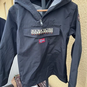Svart vindjacka från Napapijri - Svart vindjacka från Napapijri med huva och stor ficka framtill med dragkedja och kardborre. Jackan har norsk flagga broderad på bröstet och meshfoder på insidan. Perfekt för dig som gillar stilren och funktionell design. Priset är ristat i sten går ej att pruta!!