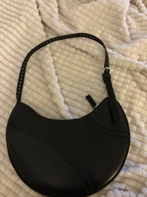 Svart axelväska i skinn med håldetaljer - Snygg svart axelväska i rundad form med coola håldetaljer på både rem och framsida. Väskan är gjord i skinn och har en dragkedja upptill. Perfekt storlek för det viktigaste och ger en stilren vibe till din outfit.