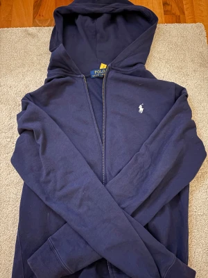 Marinblå hoodie från Polo Ralph Lauren - Snygg marinblå hoodie från Polo Ralph Lauren med klassisk vit logga på bröstet. Tröjan har huva, dragkedja och långa ärmar. Tillverkad i mjuk bomull som är skön att ha på sig. Perfekt för chill dagar och när du vill ha en clean look. Den är insydd i midjan! den är från barn avdelning alltså 170 men den är stor i modelen skulle säga som en S-M. 