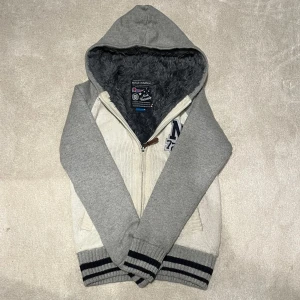 Cardigan med Pälsfoder - Säljer en mysig grå och vit hoodie med fuskpälsfoder. Den har dragkedja framtill och en broderad logga på bröstet. Perfekt för kyliga dagar med sin varma insida och ribbade muddar. Vid minsta fråga eller fundering hör av er. Byte funkar 