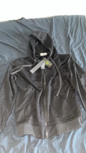 Svart hoodie från Stone Island M - Svart hoodie från Stone Island i storlek M med klassisk logopatch på ärmen. Tröjan har huva med snörning, hel dragkedja framtill och ribbade muddar. Materialet är mjuk bomull och passformen är normal. Perfekt för dig som gillar streetwear och snygga detaljer.