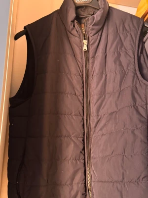  Oscar Jacobson väst - Snygg mörkblå dunväst från Oscar Jacobson med quiltad design och hög krage. Västen har dragkedja framtill och är tillverkad i lätt och slitstark polyester. Perfekt för lager-på-lager och har en stilren look som funkar året runt. Storlek 44