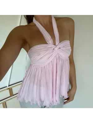 Ljusrosa omlott halterneck blus - Superfin ljusrosa blus med halterneck och omlott-detalj framtill. Toppen är plisserad och har en flowy, luftig passform som ger en söt och trendig vibe. Perfekt för dig som gillar romantiska och feminina plagg med lite extra volym.