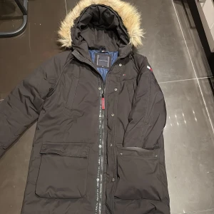 Svart parkajacka Tommy Hilfiger - Svart parkajacka från Tommy Hilfiger med huva och snygg fuskpäls. Jackan har flera fickor, dragkedja och tryckknappar framtill. Insidan är blå och jackan har klassisk Tommy Hilfiger-logga på bröstet och ärmen. Perfekt för kalla dagar och riktigt stilren. Har även spruckit under höger arm runt armhålan går knappt att märka. Ny pris ca: 1700