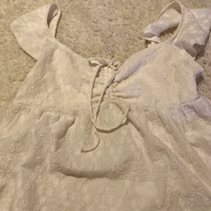 Vit topp med snöre - Supersöt vit toppg med strukturmönster och knytning framtill vid halsen. Klänningen har breda axelband och ett lätt puffigt fall. Materialet känns som bomull med en mjuk, texturerad yta och diskreta blommiga detaljer. Perfekt för sommarens och julfirande alla tillfällen!