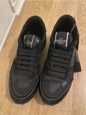 Valentino Garavani svarta sneakers - Svarta Valentino Garavani sneakers med coolt kamouflagemönster och platt sula. Skorna har snörning och en rund tå, samt detaljer i både skinn och syntet. Märkeslogga på plösen och en urban vibe som passar perfekt till streetwear.