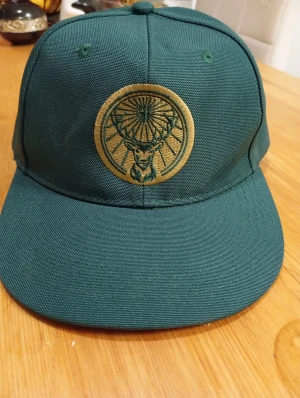 Grön Jägermeister keps med broderi - Snygg grön keps från Jägermeister med gul broderad logga med hjort på framsidan. Märket syns även på en orange etikett på sidan. Kepsen har klassisk form och är tillverkad i slitstark bomull. Perfekt för dig som vill sticka ut med en unik accessoar.