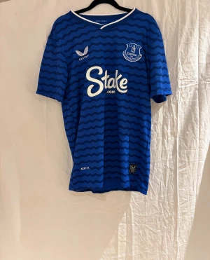 Everton 25/26 Grealish 18 - Snygg blå Everton fotbollströja med vågigt mönster, klubbmärke på bröstet och sponsortryck. Nummer 18 och namnet Grealish på ryggen. Tillverkad i lätt och ventilerande funktionsmaterial, perfekt för match eller träning.
