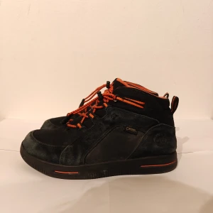 Timberland kängor  - Säljer ett par svarta Timberland sneakers i mocka och textil med orange snören och detaljer. Skorna har rund tå, platt sula och snörning. Gore-Tex-märke på sidan för extra väderskydd. Perfekta för dig som gillar streetstyle och funktion.