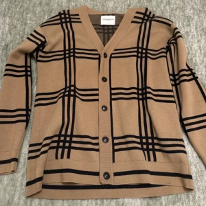 Beige rutig kofta från These Glory Days - Snygg beige kofta med svart rutigt mönster från These Glory Days. Koftan har v-ringad hals, knappar framtill och långa ärmar. Materialet är mjukt och känns skönt mot huden, perfekt för lager-på-lager. Passar dig som gillar klassisk stil med en modern twist.