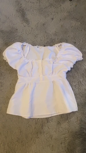 Vit blus med spets och puffärm - Säljer en söt vit blus med puffiga korta ärmar och spetsdetaljer längs halsringningen och ärmsluten. Blusen har knappar framtill och en markerad midja som ger en feminin siluett. Perfekt för dig som gillar romantisk stil.