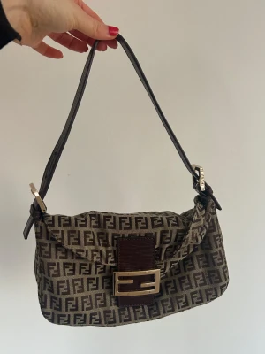 Fendi axelväska  - AxelväKa från Fendi med FF-logomönster i brunt och beige. Väskan har ett justerbart axelband i mörkbrunt skinn, guldfärgat spänne och detaljer i skinn. Insidan har dragkedja och Fendi-logga på metalldetaljer. Knappt använd, köpt på Collectorscage i Köpenhamn som har äkthetsgaranti för ca 10 000 kr.