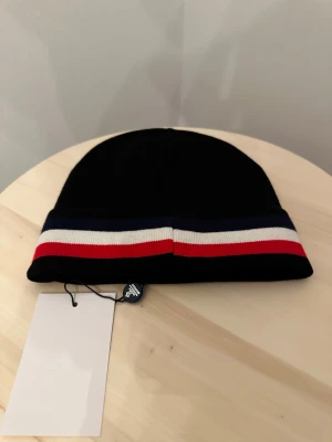 Svart Moncler mössa med ränder - Svart mössa Moncler med klassiska blå, vita och röda ränder runt kanten. Mössan har Moncler-logga framtill och är tillverkad i mjukt material som passar perfekt för kyliga dagar. Snygg och stilren accessoar för dig som gillar exklusiva märken.