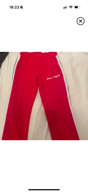 Palm Angels röda mjukisbyxor S - Palm Angels röda mjukisbyxor i storlek S med vita sidoränder och logotyp på benet. Byxorna har elastisk midja och är tillverkade i 100% polyester för en skön och sportig vibe. Perfekt för dig som gillar streetwear och vill sticka ut.