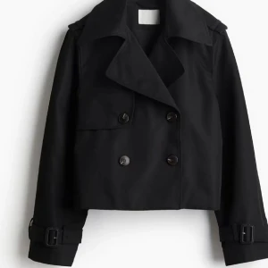 Svart croppad trenchcoat från h&m - Snygg svart croppad trenchcoat med bred krage och dubbla knapprader. Jackan har långa ärmar med spännen vid mudden och en cool, boxig passform. Perfekt för dig som gillar stilrena och trendiga plagg med edge.