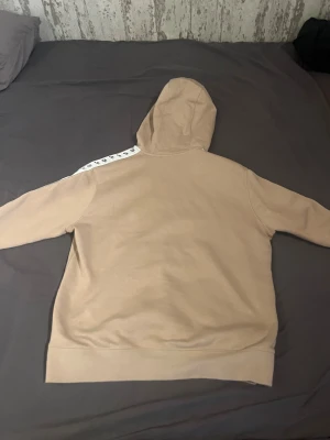 Beige Nike hoodie med logoband - Säljer en beige hoodie från Nike med svart snörning i huvan och vita band med svarta Nike-loggor längs ärmarna. Tröjan har en klassisk känguruficka framtill och ribbade muddar vid ärmslut och nederkant. Perfekt för chill eller streetwear.