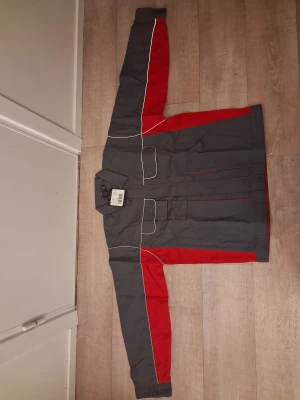 Grå och röd twilljacka från Univern - Säljer en grå och röd twilljacka från Univern i storlek XL. Jackan har vita kontrastsömmar, flera fickor med lock framtill och klassisk krage. Materialet är slitstark bomull och polyester, perfekt för dig som gillar funktionell och snygg workwear.