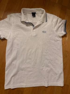 Vit pikétröja från Hugo Boss - Vit pikétröja från Hugo Boss med klassisk krage och korta ärmar. Tröjan har subtilt blått Boss-logo broderat på bröstet och blå detaljer på krage och ärmslut. Regular fit och tillverkad i mjuk bomull som andas. Perfekt för en clean och fräsch look.