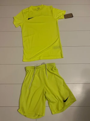 Neongula träningsshorts och T-shirt från Nike - Säljer ett par neongula Nike Dri-Fit shorts och T-shirt med vit snörning i midjan och svart Nike-logga på benet. De är tillverkade i lätt polyester som andas och har en loose passform, perfekta för sport eller gym. Midjan är elastisk och materialet är mjukt mot huden.