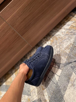 Mörkblå Christian Louboutin sneakers - Snygga mörkblå sneakers från Christian Louboutin i mocka med klassisk röd sula och signaturtext undertill. Skorna har rund tå, platt sula och snörning framtill. Insidan är beige och de har en stilren, modern look som sticker ut med den ikoniska röda undersidan.