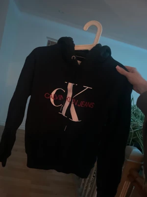 Svart hoodie Calvin Klein Jeans XS - Svart hoodie från Calvin Klein Jeans i storlek XS. Stor CK-logga i vitt och rött på bröstet, med snörning i huvan. Mjuk och skön bomullskvalitet, perfekt för chill dagar. Klassisk passform med långa ärmar och ribbade muddar.