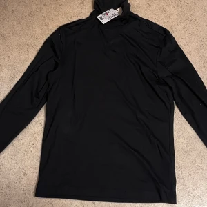 Svart polotröja slim fit från H&M - Svart polotröja från H&M i slim fit-modell. Tröjan har lång ärm och hög krage som ger en clean och stilren look. Materialet är mjukt och stretchigt, perfekt för lager-på-lager under höst och vinter. Passformen är tight och modern.